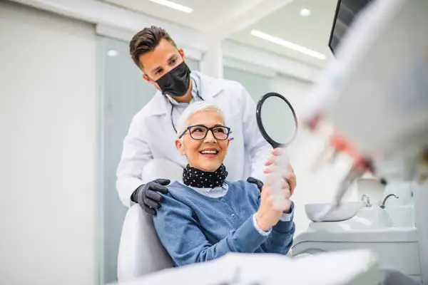 Beautiful senior woman smiling and holding up a mirror to her dental implants at Mountain State Oral and Facial Surgery in Parkersburg, WV Beautiful senior woman smiling and holding up a mirror to her dental implants at Mountain State Oral and Facial Surgery in Parkersburg, WV